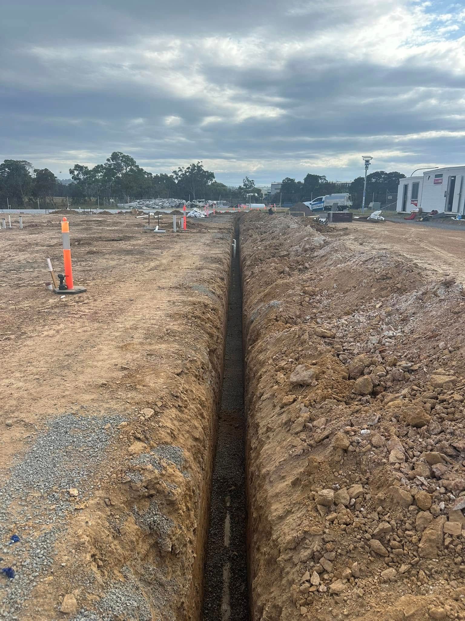 Long trench cut through a dirt construction site under a cloudy sky, with orange markers nearby.