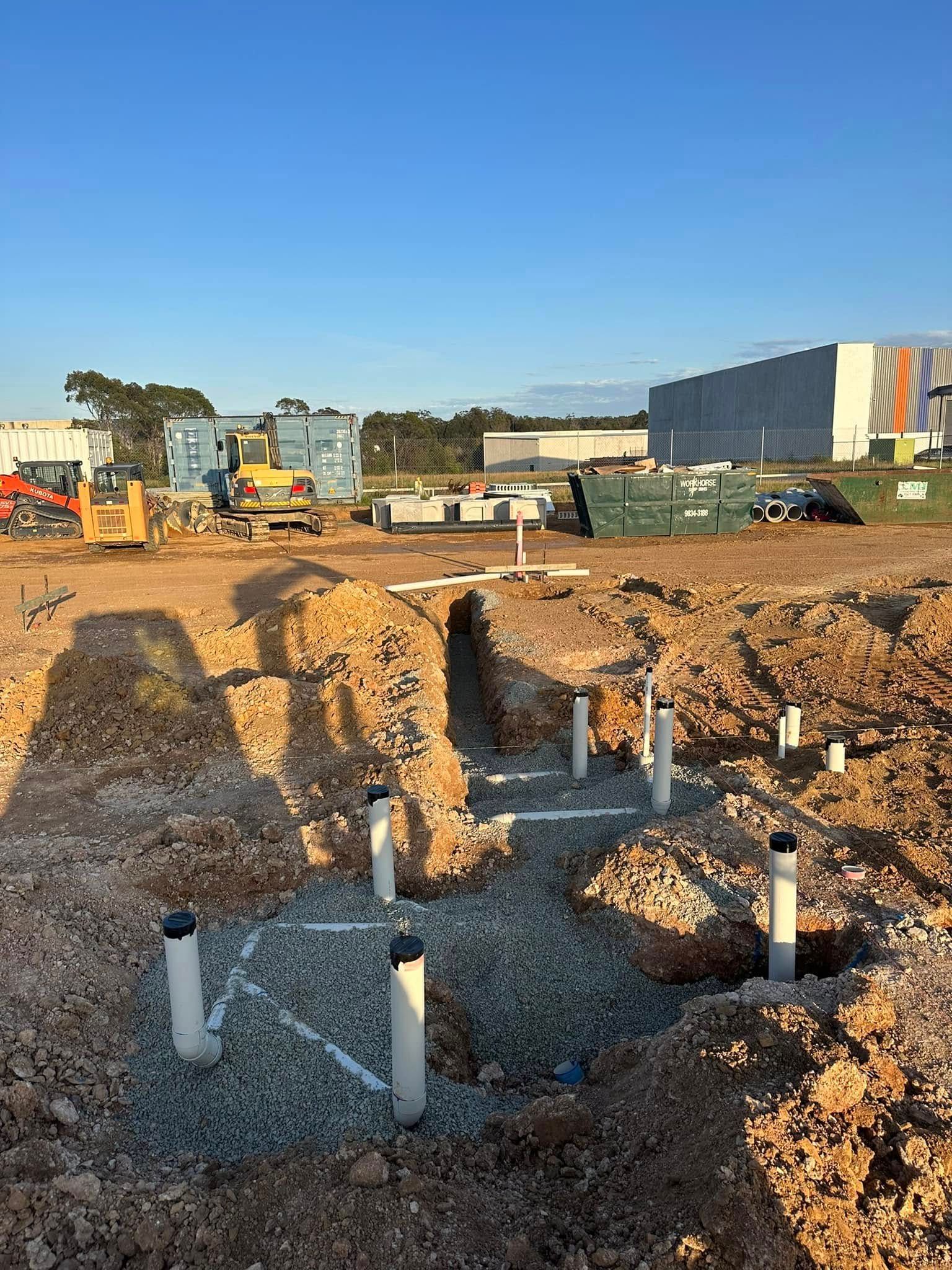 Construction site with excavated trench, exposed pipes, gravel, and heavy equipment under a clear blue sky