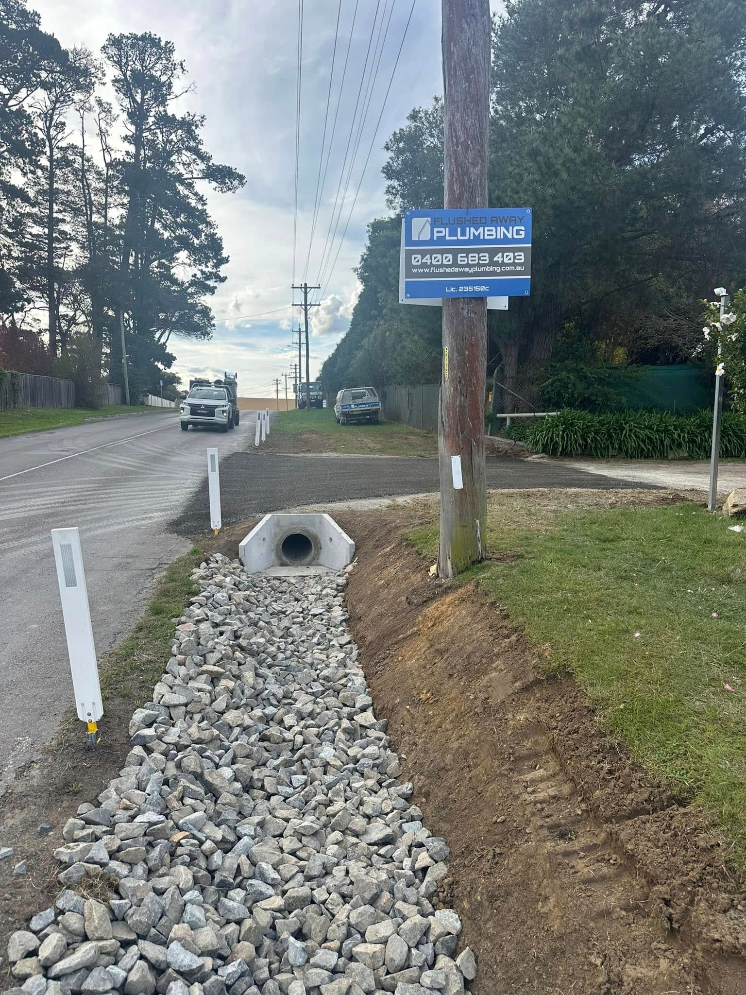 Roadside drainage ditch with rocks, concrete culvert, and a blue sign beside a quiet rural road.
