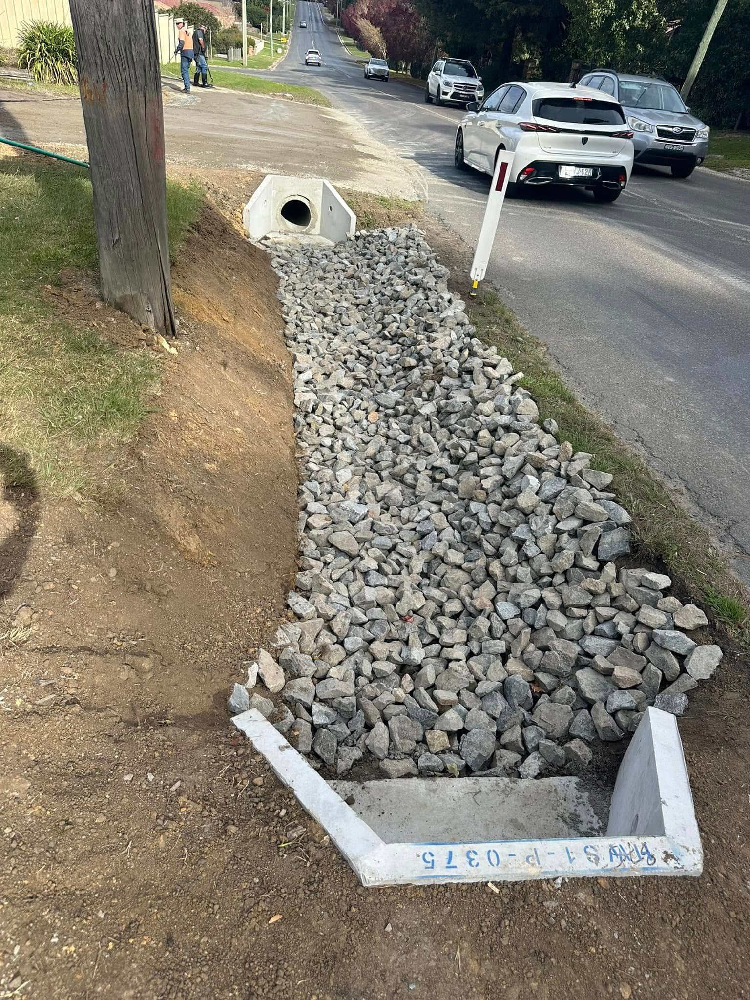 Rock-filled drainage trench beside a road, with white concrete borders and a drain opening at the top