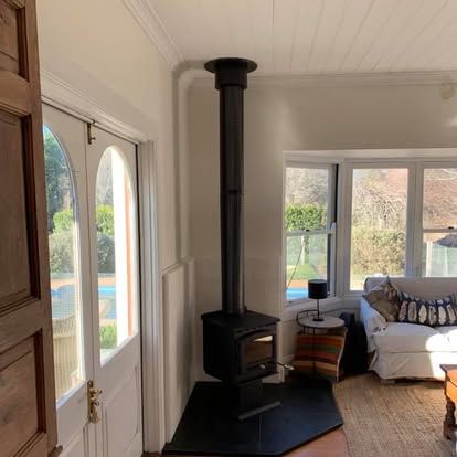 Bright living room with a black wood stove, sofa, and large windows overlooking a garden.