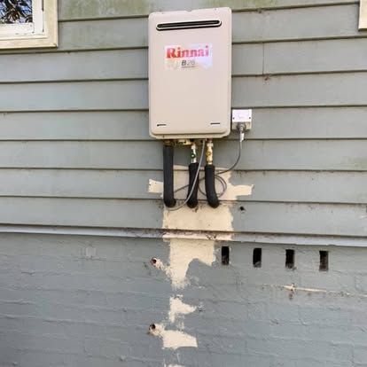Rinnai tankless water heater mounted on blue house siding with exposed pipes and patchy paint repairs