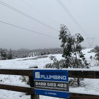 Snow-covered roadside with a plumbing sign in the foreground and power lines over a foggy landscape.