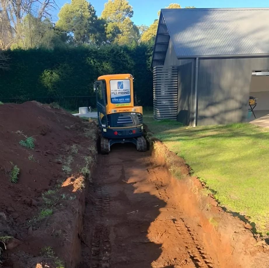 Mini excavator digging a trench beside a gray building and hedge on a sunny day