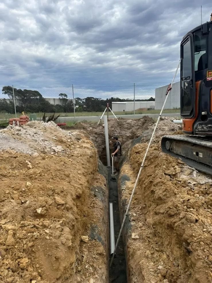 Excavator beside a deep trench at a construction site, with soil piles and support braces.