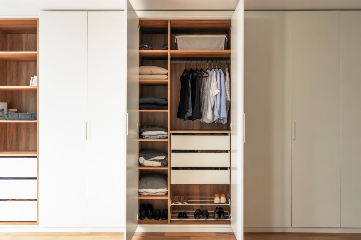 White and wood-toned walk-in closet with doors open, showcasing clothes, drawers, and shelving.