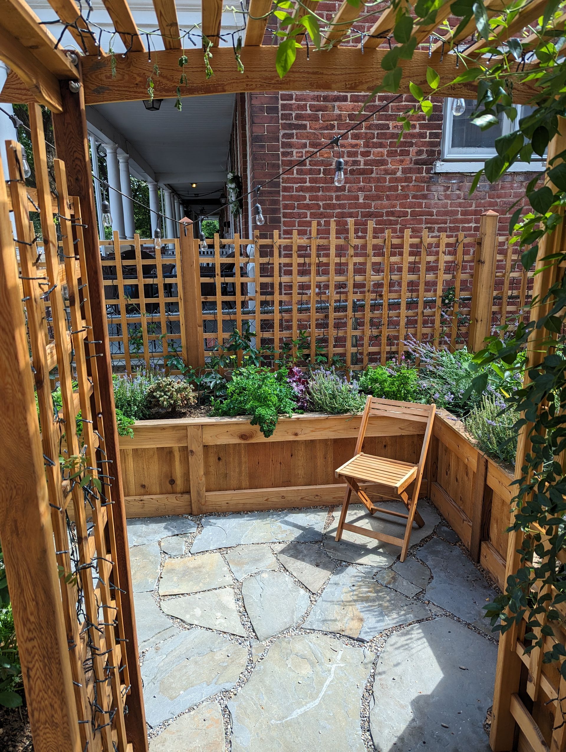 Wooden pergola frames a small patio with a chair, flower beds, and a brick house.
