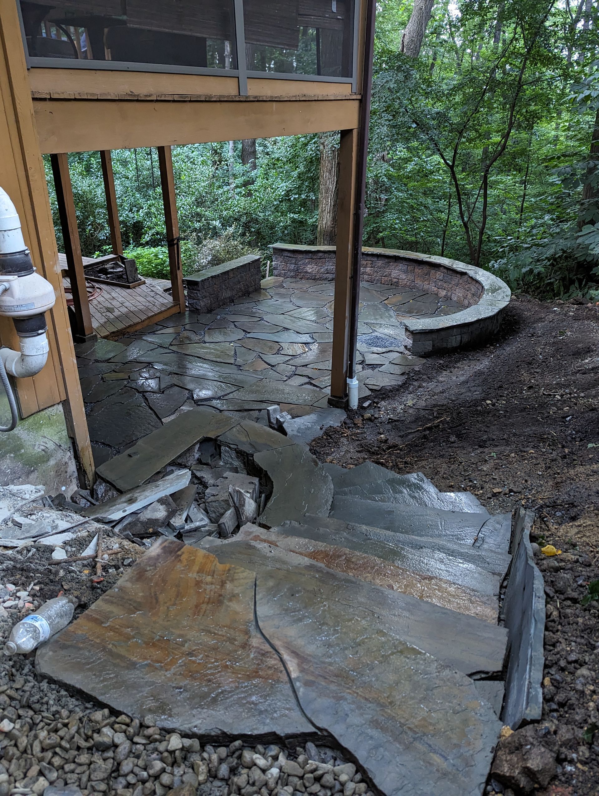 Outdoor patio with stone floor and curved retaining wall near a wooden structure and trees.