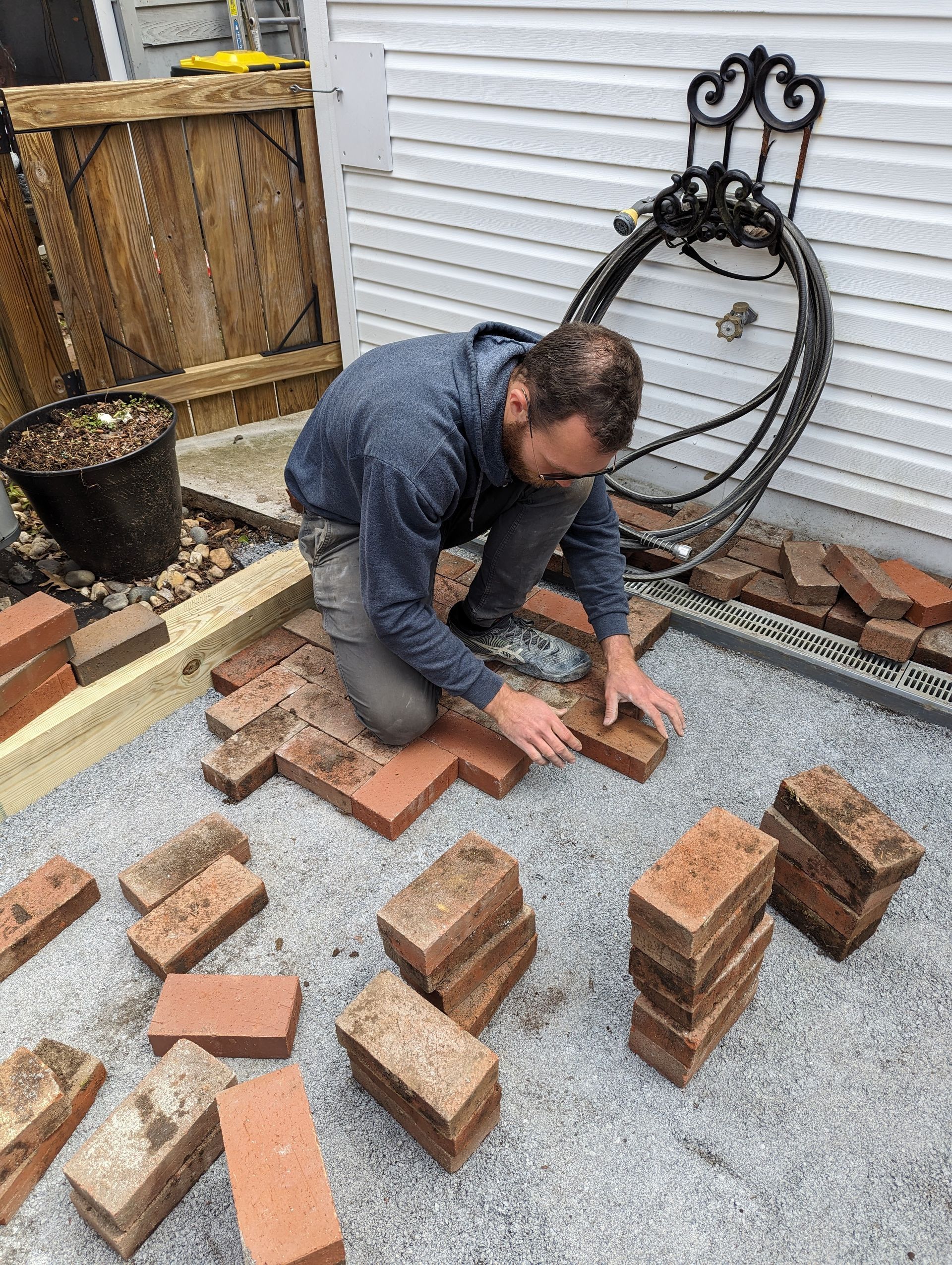 Man laying red bricks on a gravel patio. He's arranging them to create a pattern next to a building.