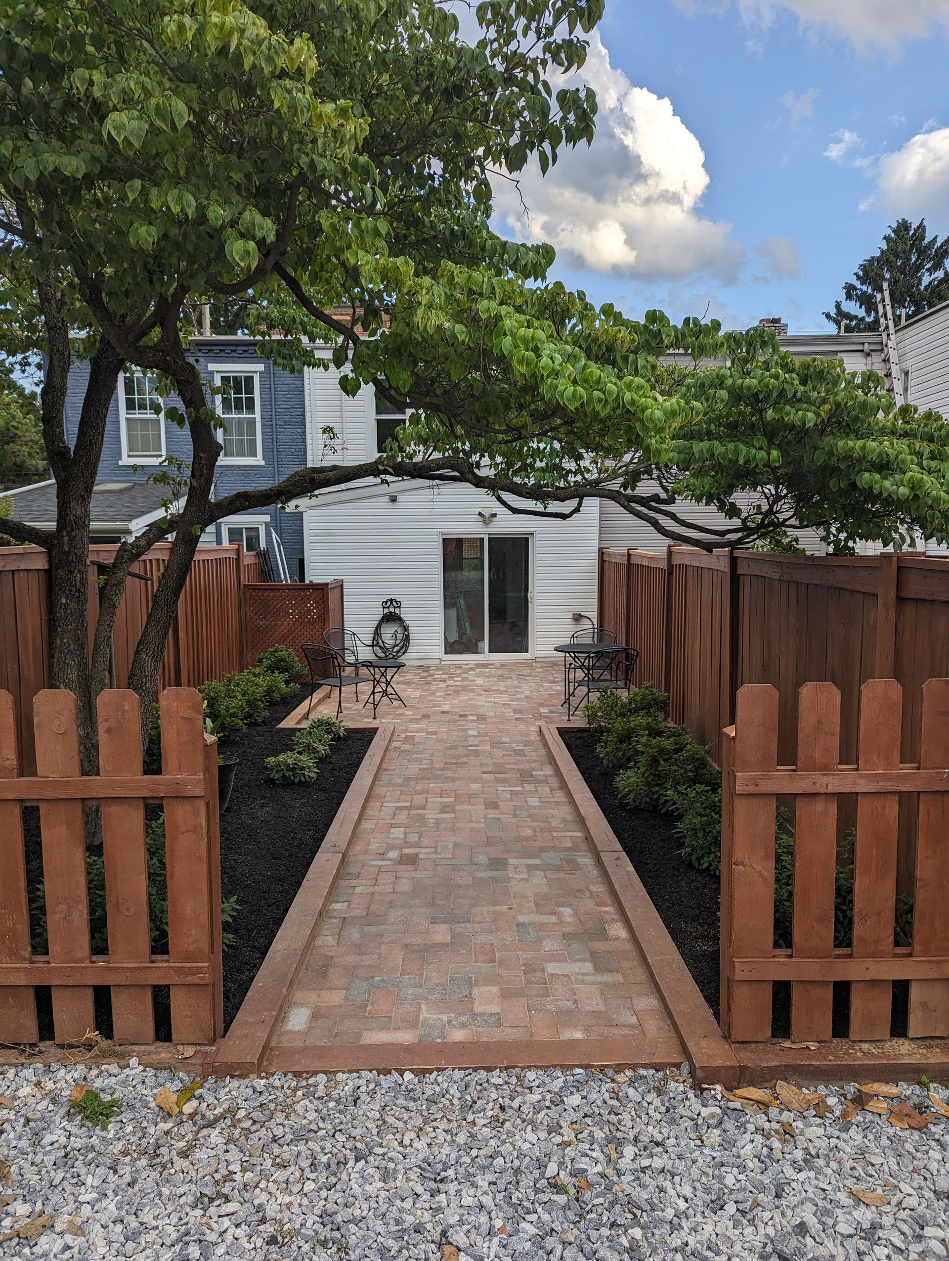 Brick pathway leads to sliding glass door, enclosed by brown wooden fence and lush greenery.