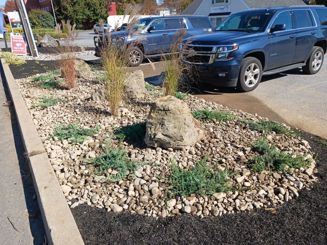 Landscaped bed with rocks, mulch, and greenery; two blue SUVs parked nearby.