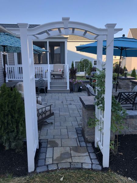 White arbor frames a paved pathway leading to a screened porch, with outdoor seating and umbrellas.