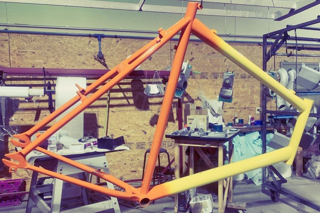 Orange and yellow bike frame hanging in a workshop, partially painted.