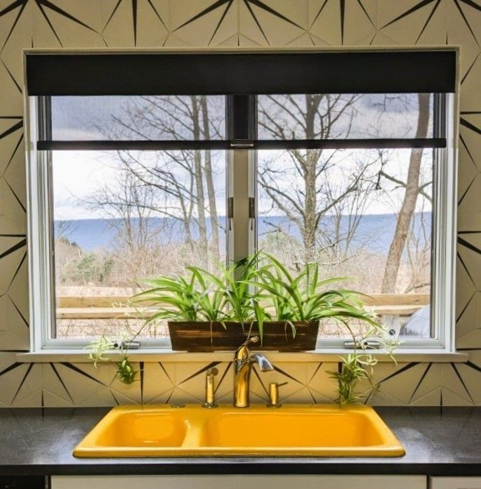 Yellow sink in a kitchen with a view of trees and mountains, plants on the windowsill, black window shade.