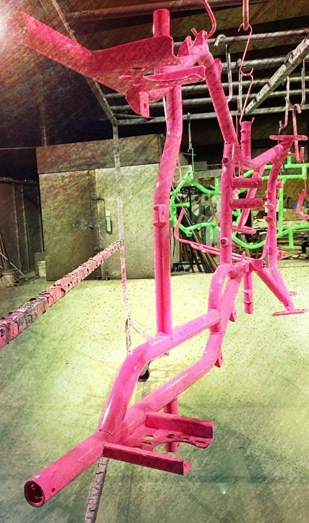 Pink metal frame of a bicycle part hanging on a production line; green frame visible in the background.