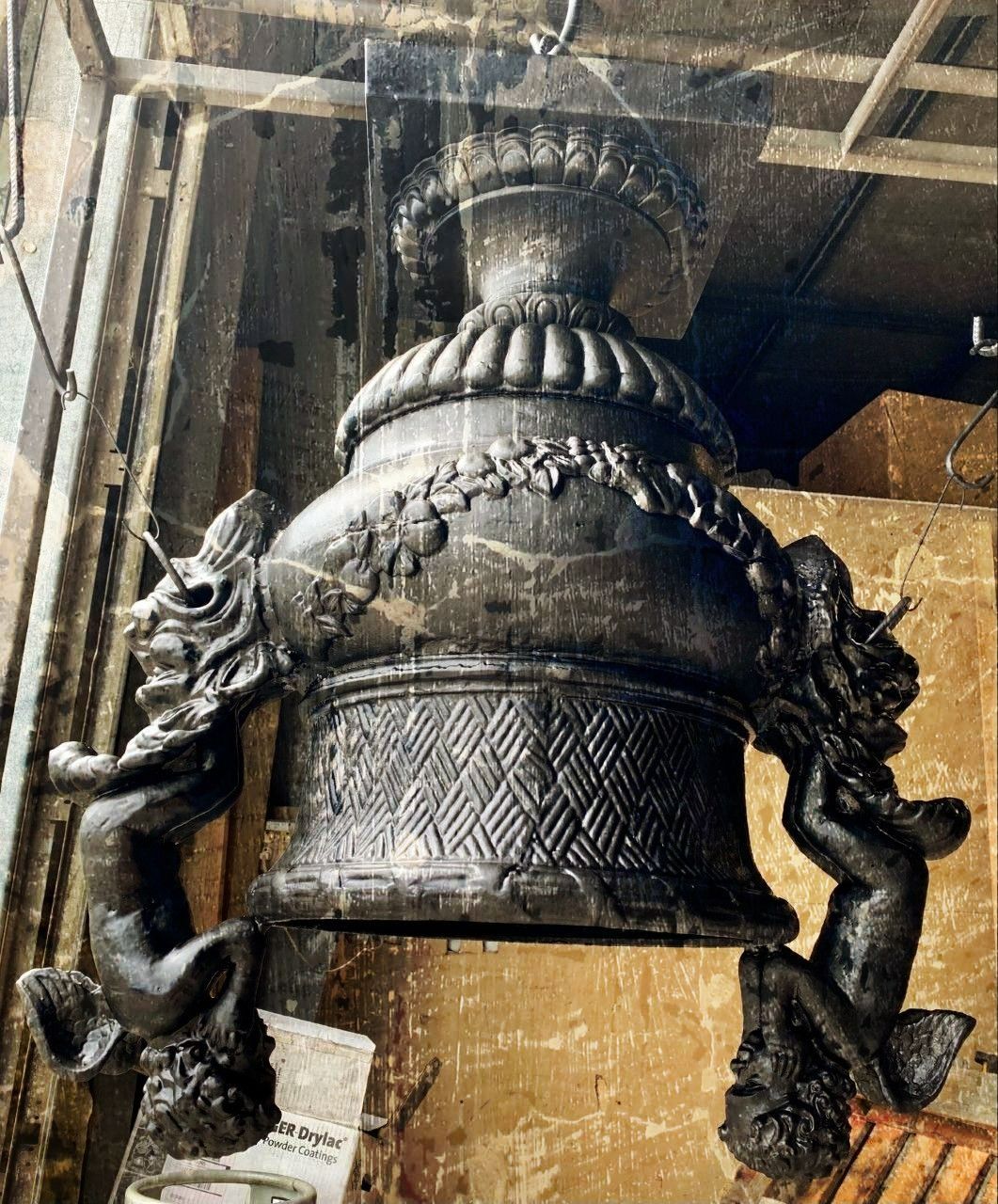 Black, ornate urn with cherub-like figures on either side. It is in a workshop.