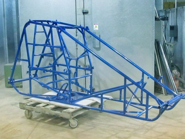Blue race car frame on a wooden pallet inside a gray industrial setting.
