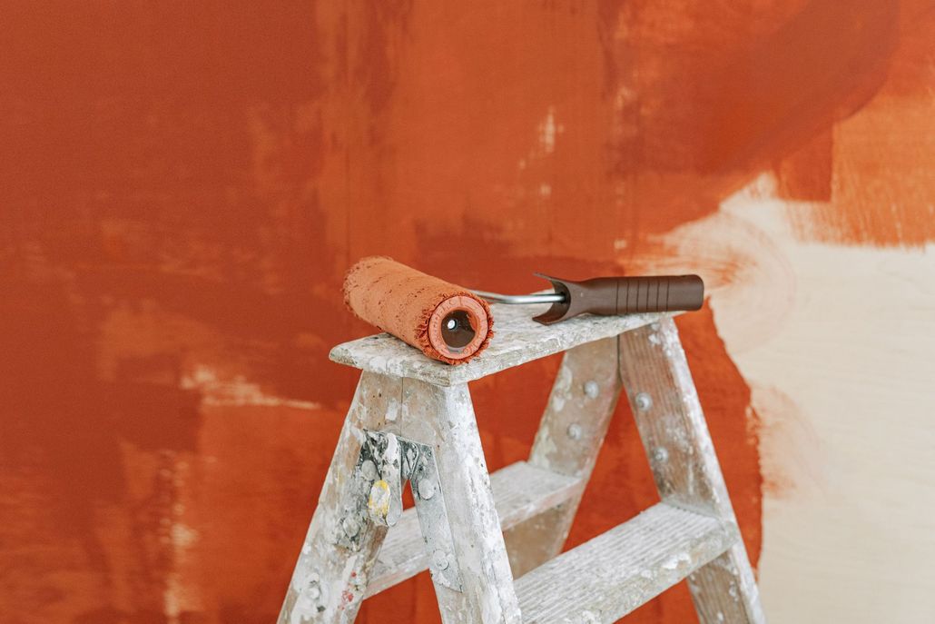 Paint roller resting on a stepladder against a partially painted orange wall