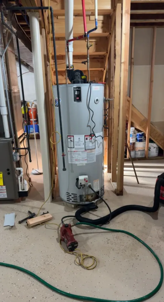 Water heater in an unfinished basement with exposed framing, hoses, and tools on the floor