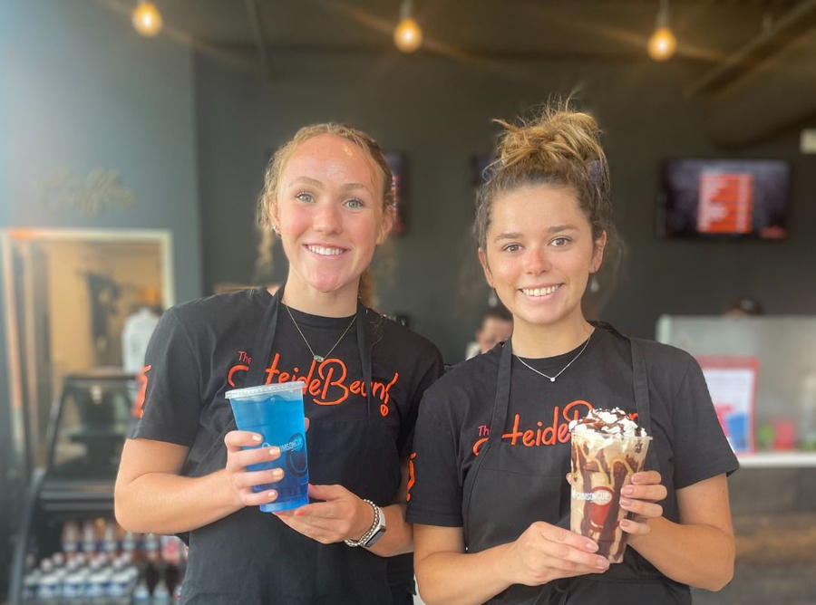 Two women in black aprons holding colorful drinks, smiling in a cafe setting.