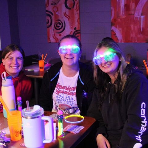 Three people smiling at a table with glow-in-the-dark glasses, beverage containers.