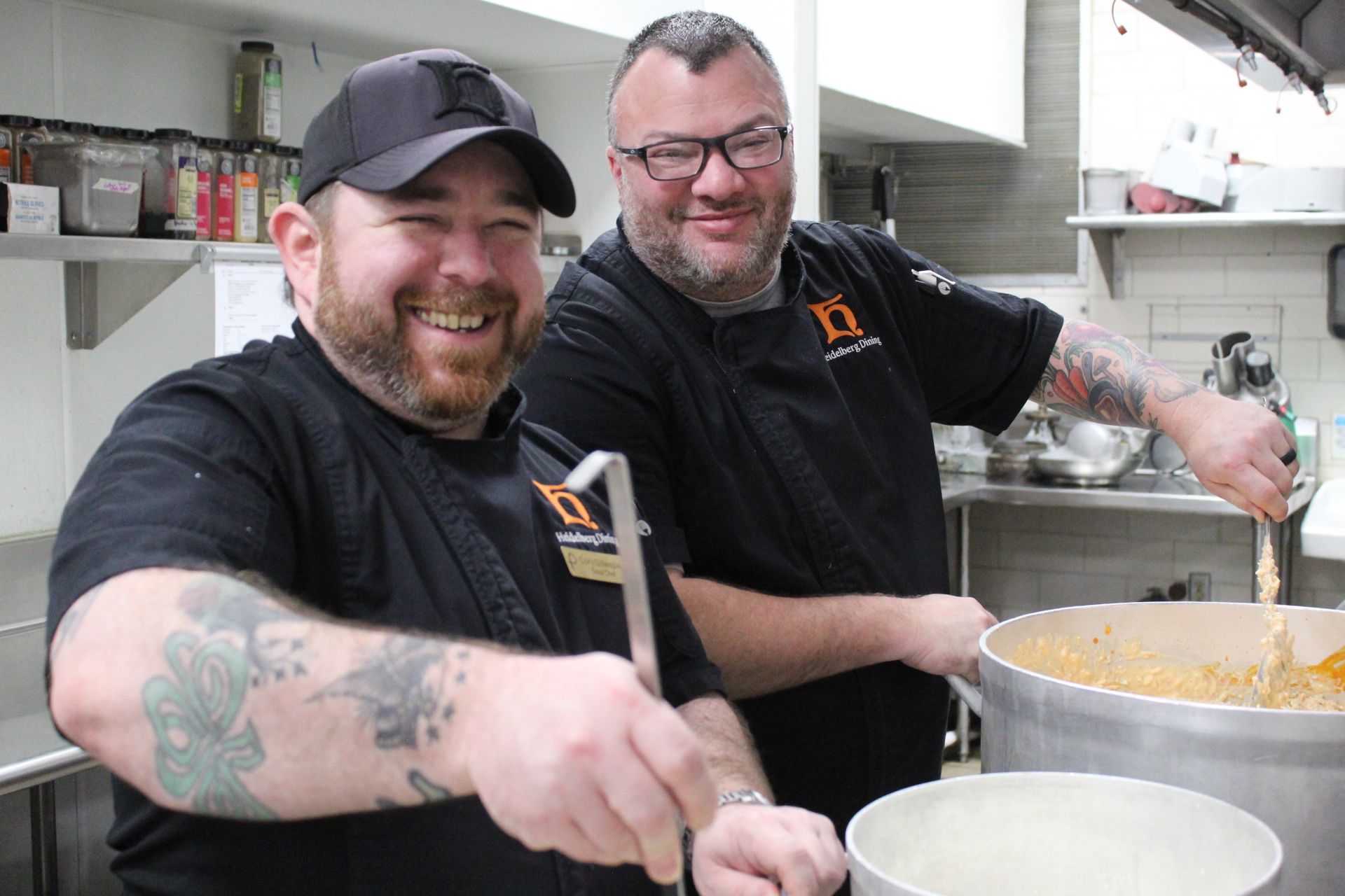 2 Chefs stirring food in a pan on a stove in a commercial kitchen.