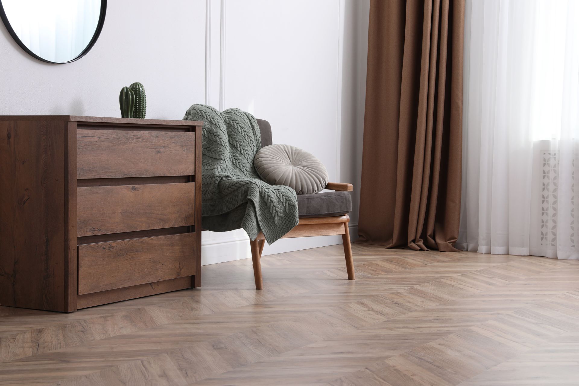 Wooden dresser, armchair with blanket and pillow, and brown curtains in a well-lit room.
