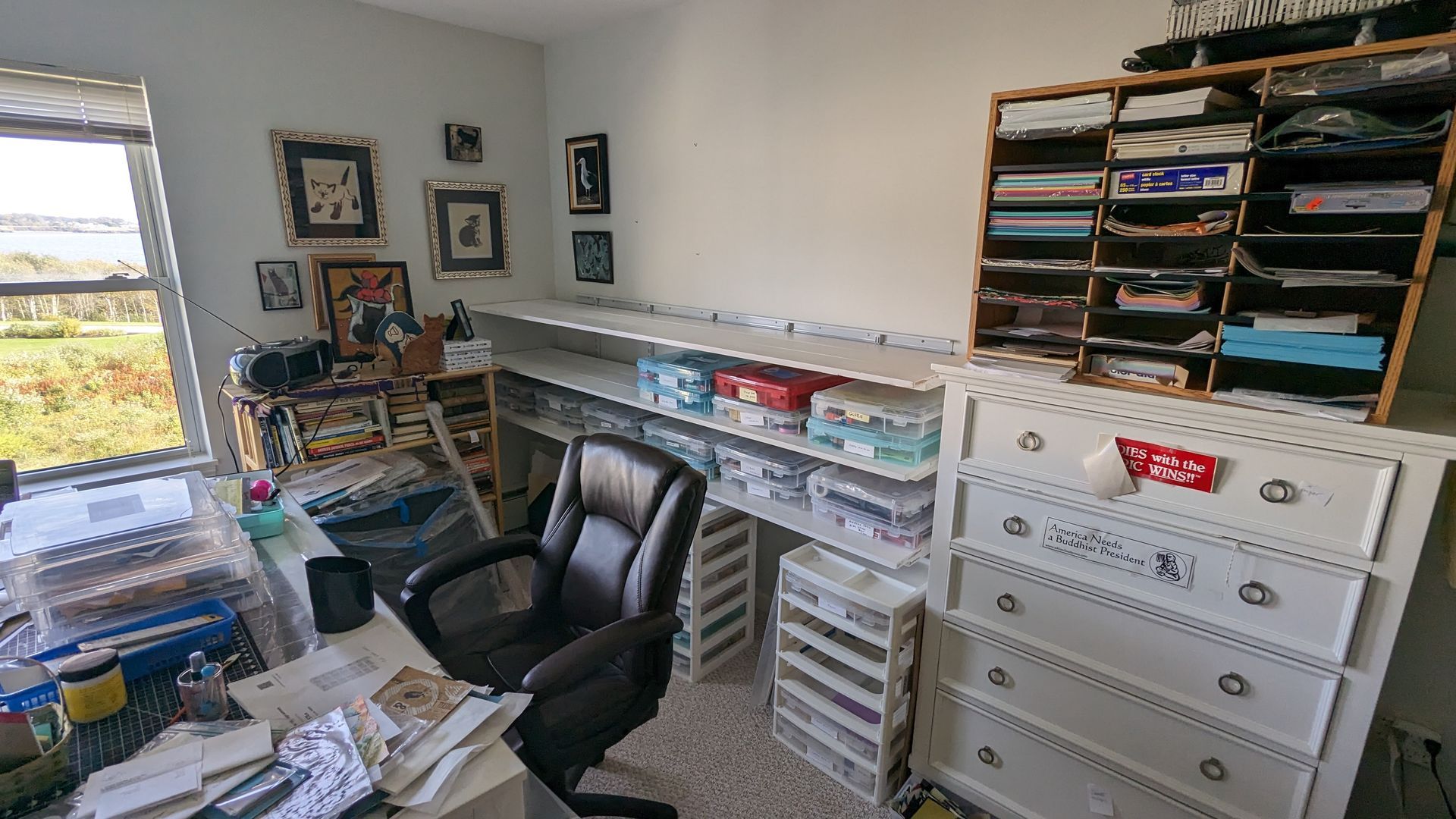 A room with a desk , chair , dressers , shelves and a window.