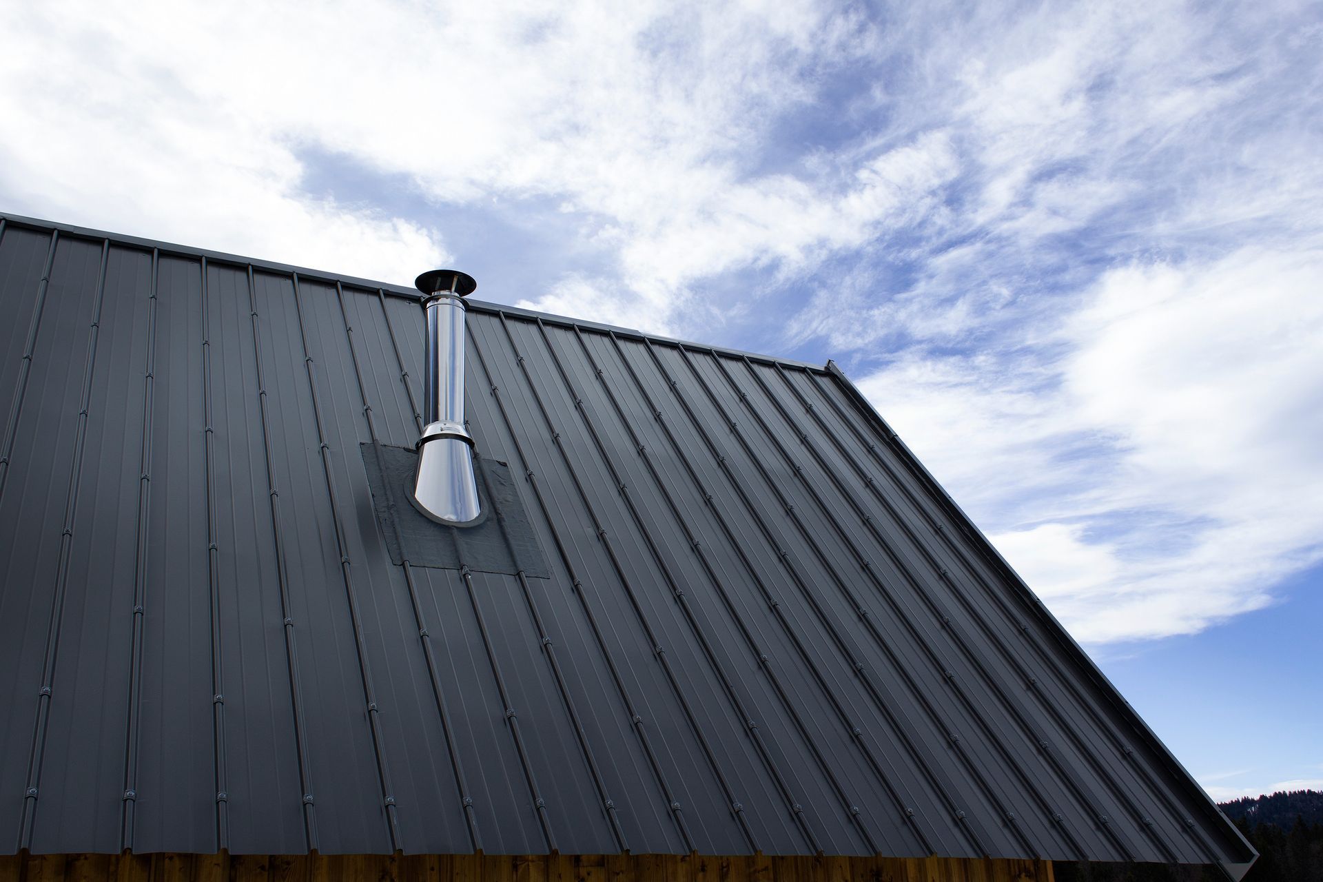 Dark metal roof with a silver chimney pipe against a cloudy blue sky