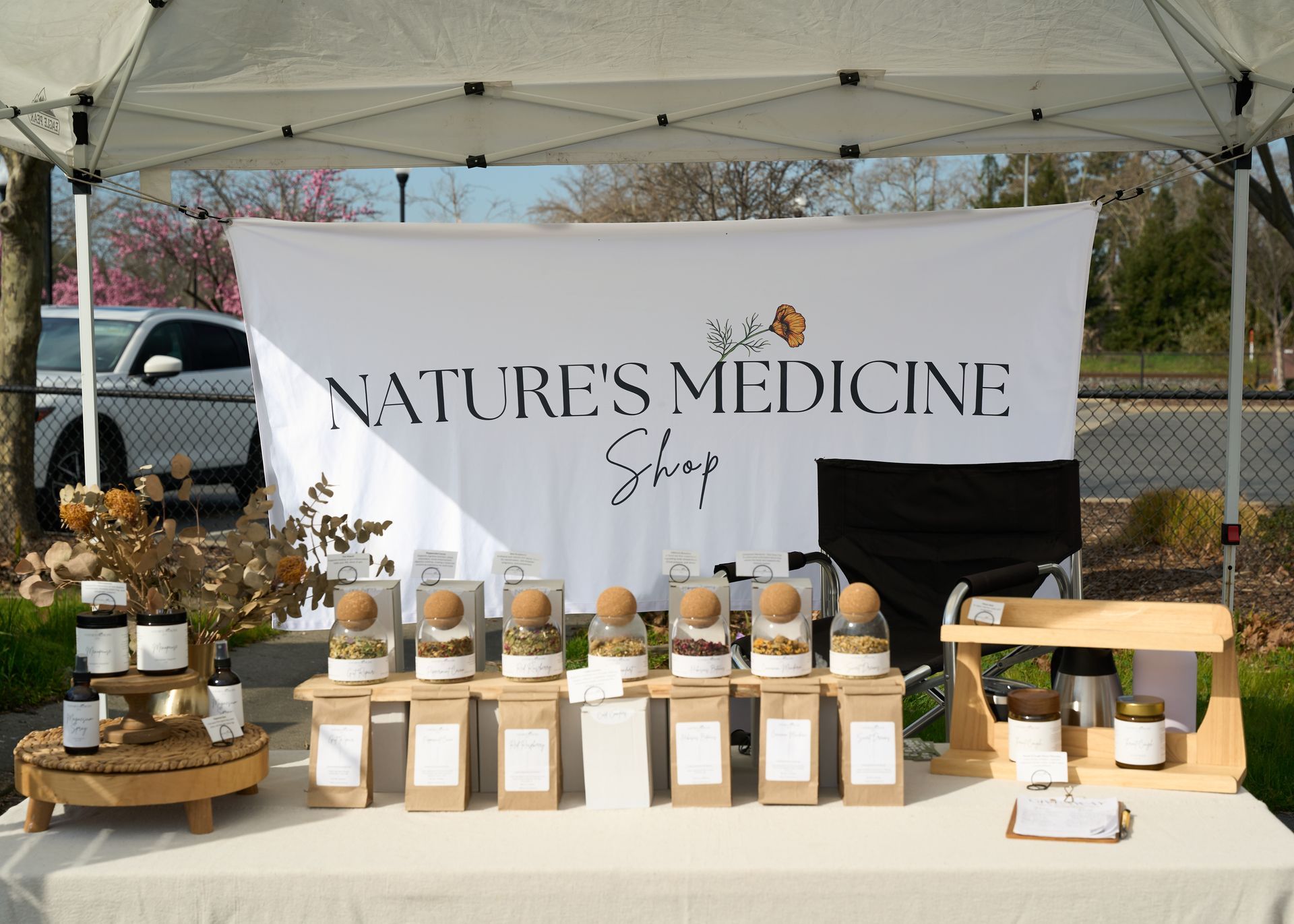 Vendor booth featuring a Nature's Medicine banner, jars of herbal products, and fresh flowers under a white canopy at the Loomis Community Farmers Market
