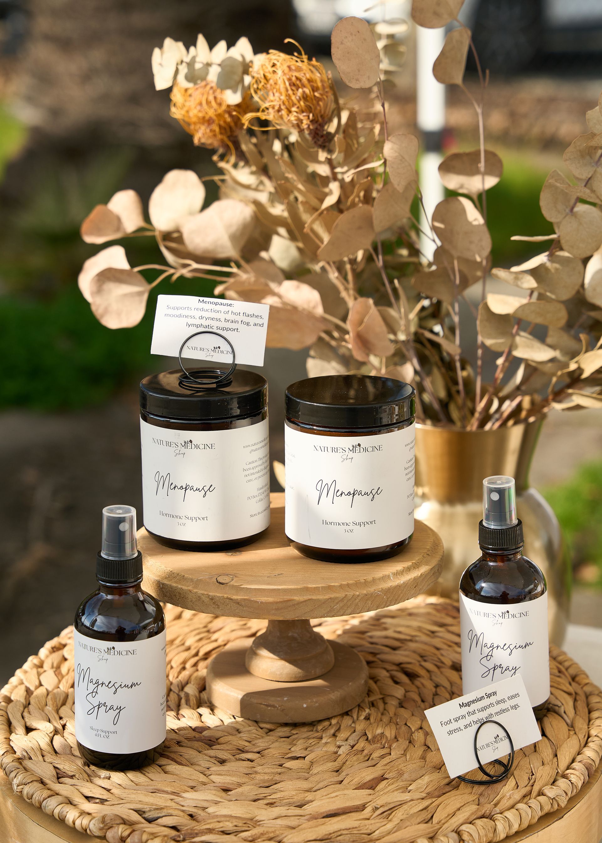 Rustic display of black-labeled wellness jars and bottles with dried beige flowers on woven trays and mats at the Loomis Community Farmers Market