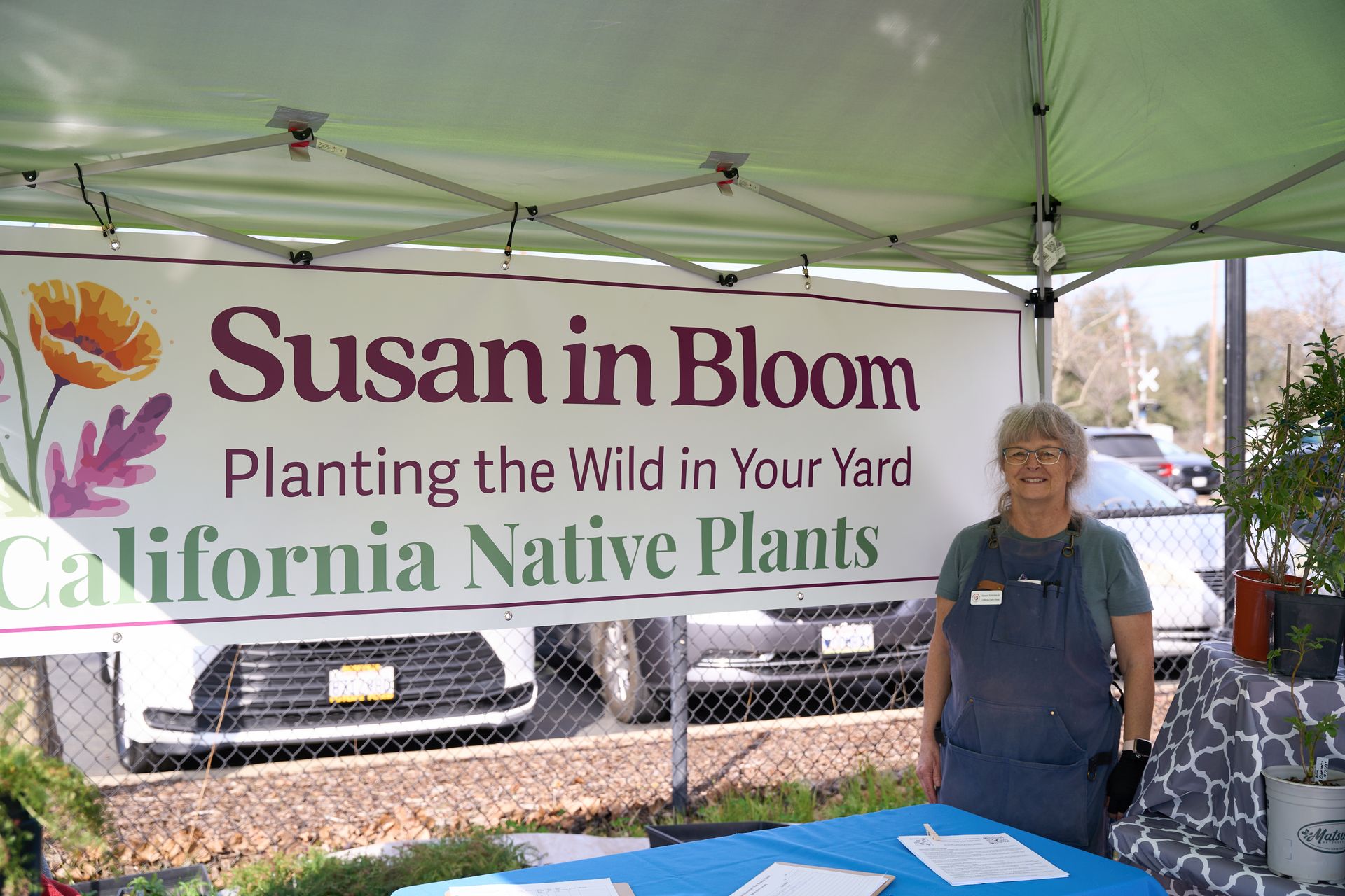 Susan in Bloom plant nursery booth under a green canopy, with a woman standing beside a table.