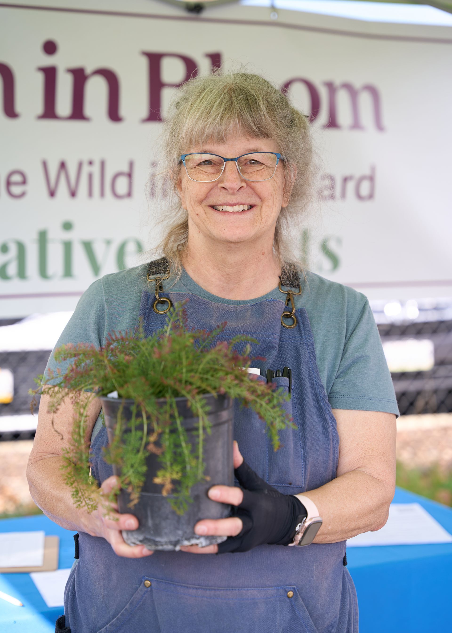 Woman holding a potted fern at a plant vendor booth with a branded backdrop banner at the Loomis Community Farmers Market