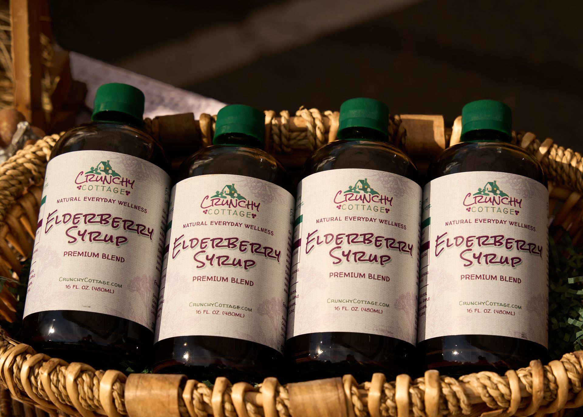 Four bottles of handcrafted elderberry syrup with green caps and cream labels displayed in a woven basket at the Loomis Community Farmers Market