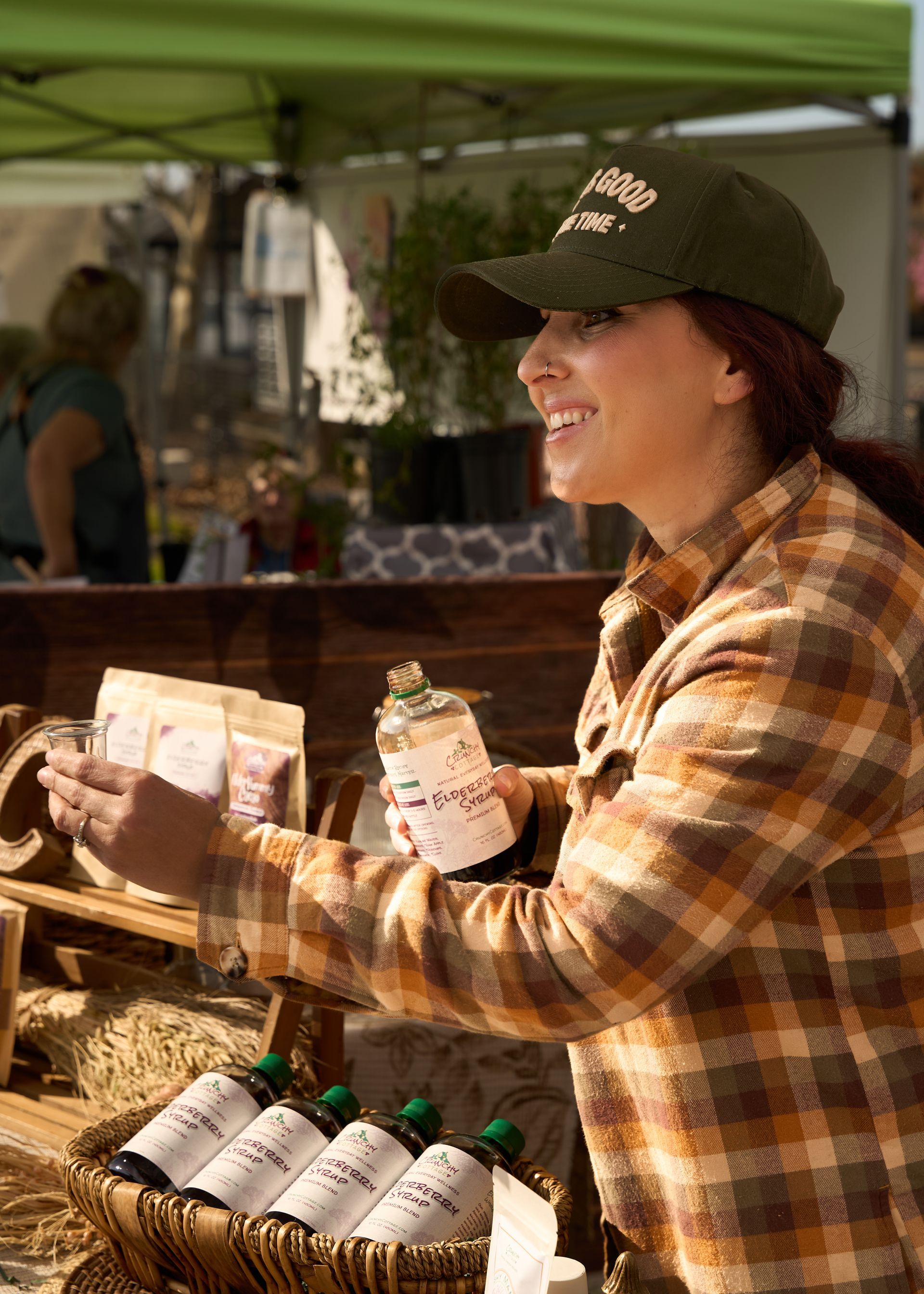 Vendor in a plaid shirt and cap handing out cold bottled drinks at an outdoor market stall during the Loomis Community Farmers Market