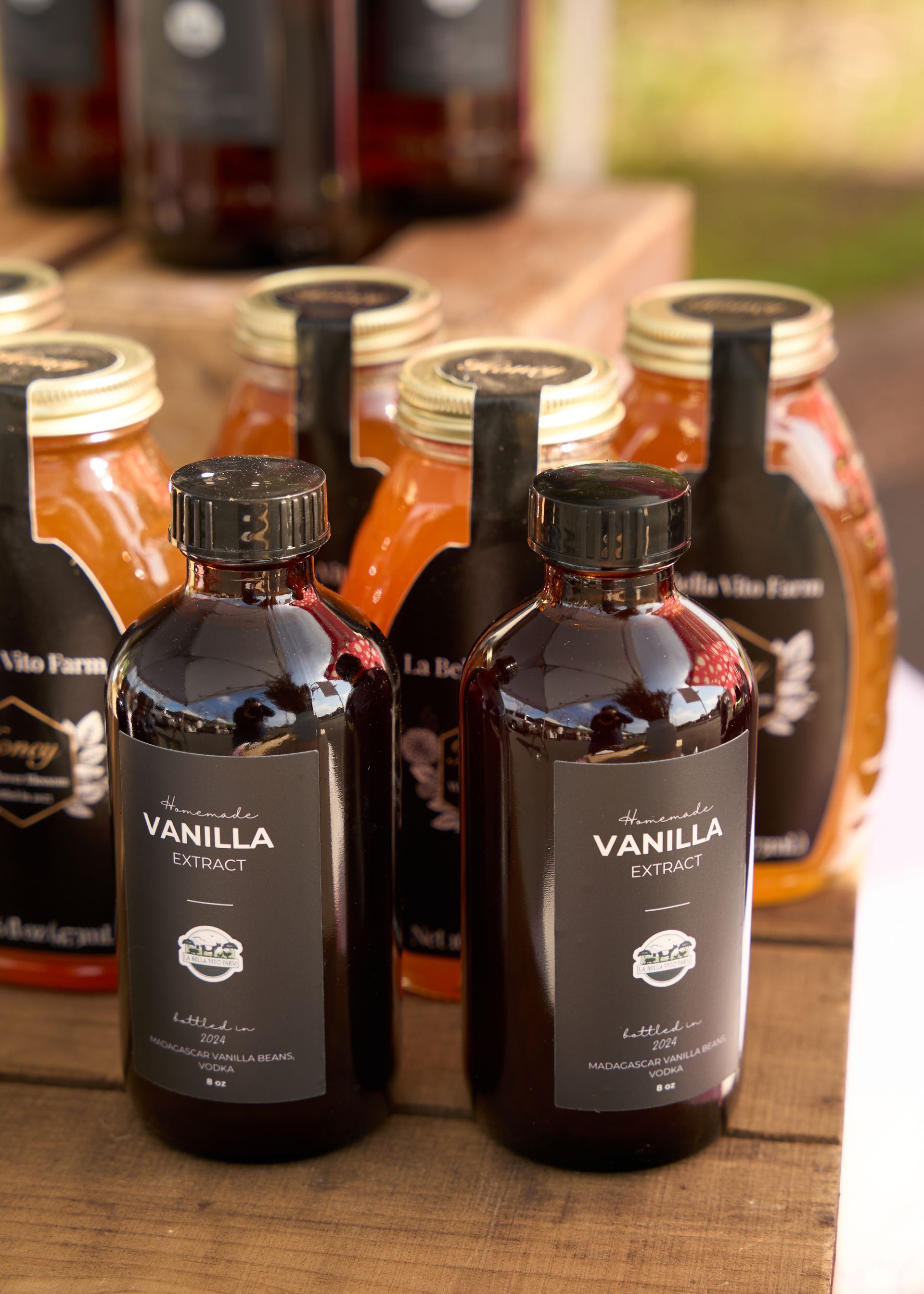 Dark amber vanilla bottles on a rustic wooden display table with blurred jars and bottles at the Loomis Community Farmers Market