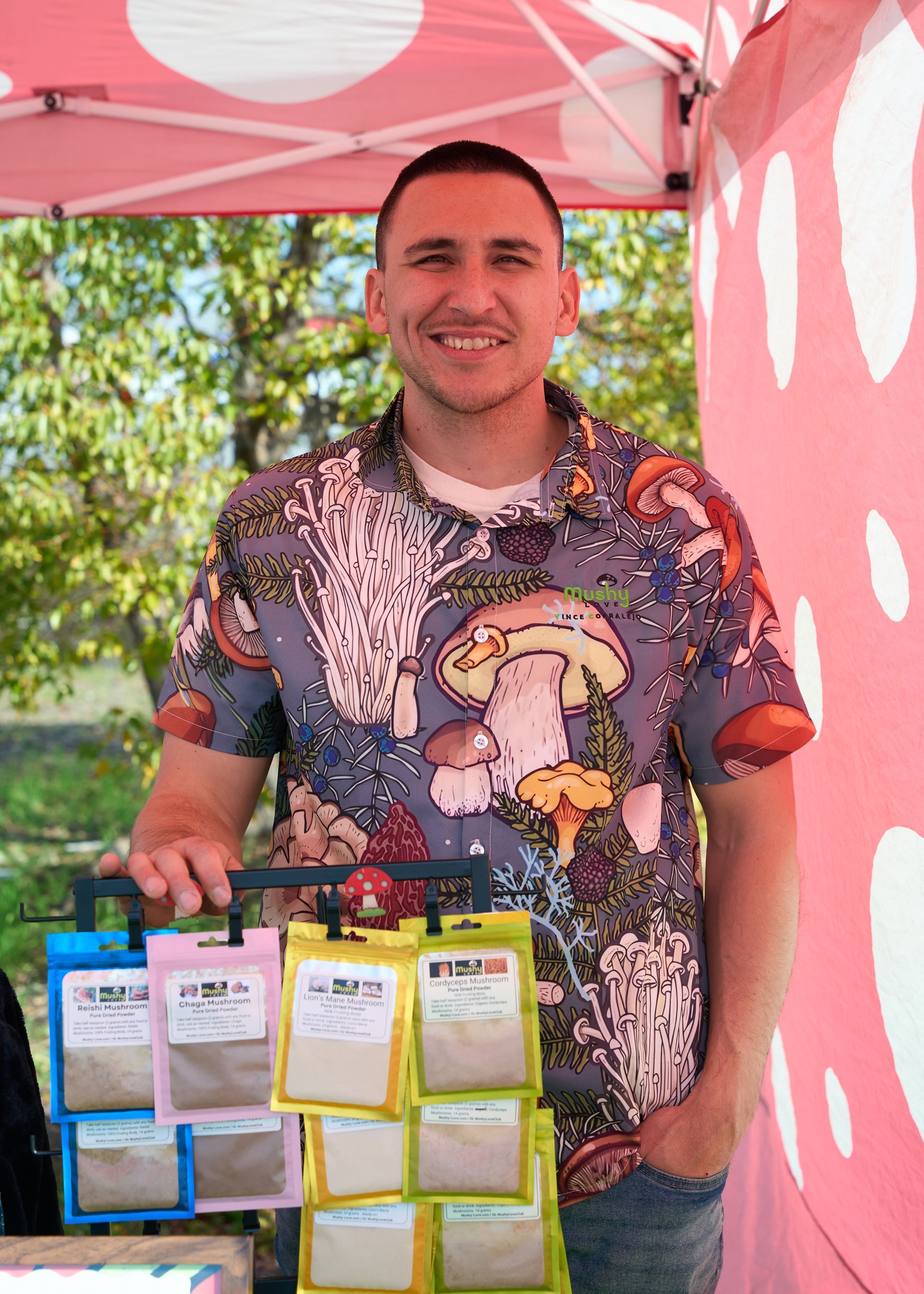 Smiling vendor standing behind a display of colorful packaged artisan goods under a pink canopy at the Loomis Community Farmers Market