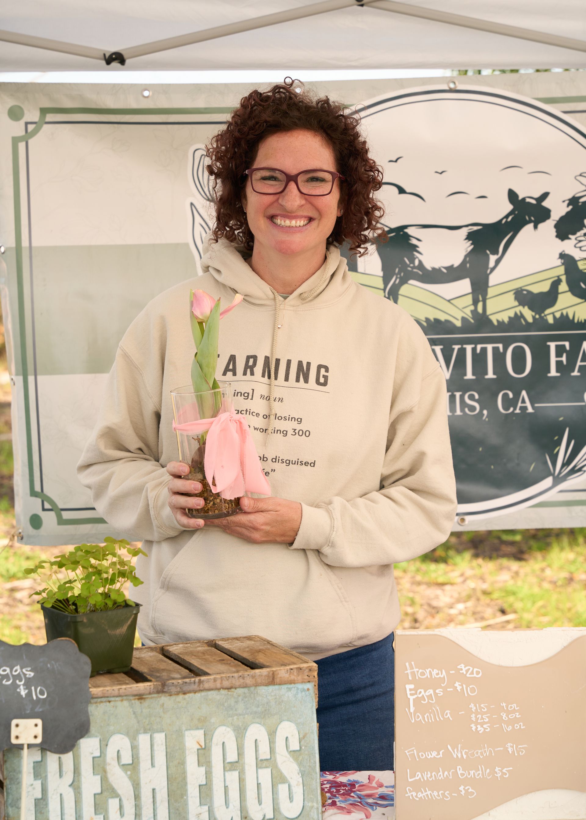 A smiling woman holding a pink flower at a busy farm stand at the Loomis Community Farmers Market