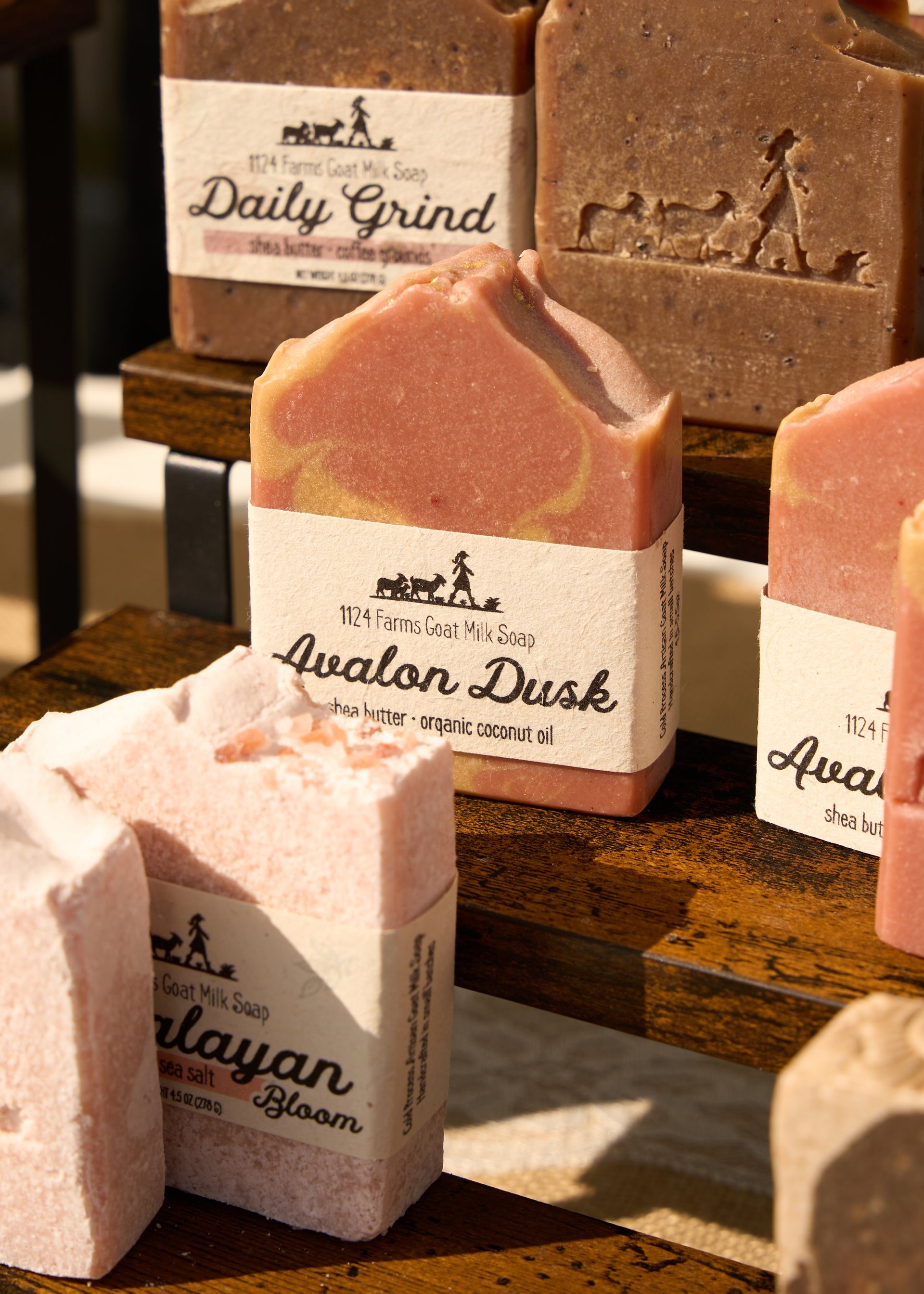 Assorted handmade pink and brown soap bars displayed on wooden shelves with scent labels at the Loomis Community Farmers Market