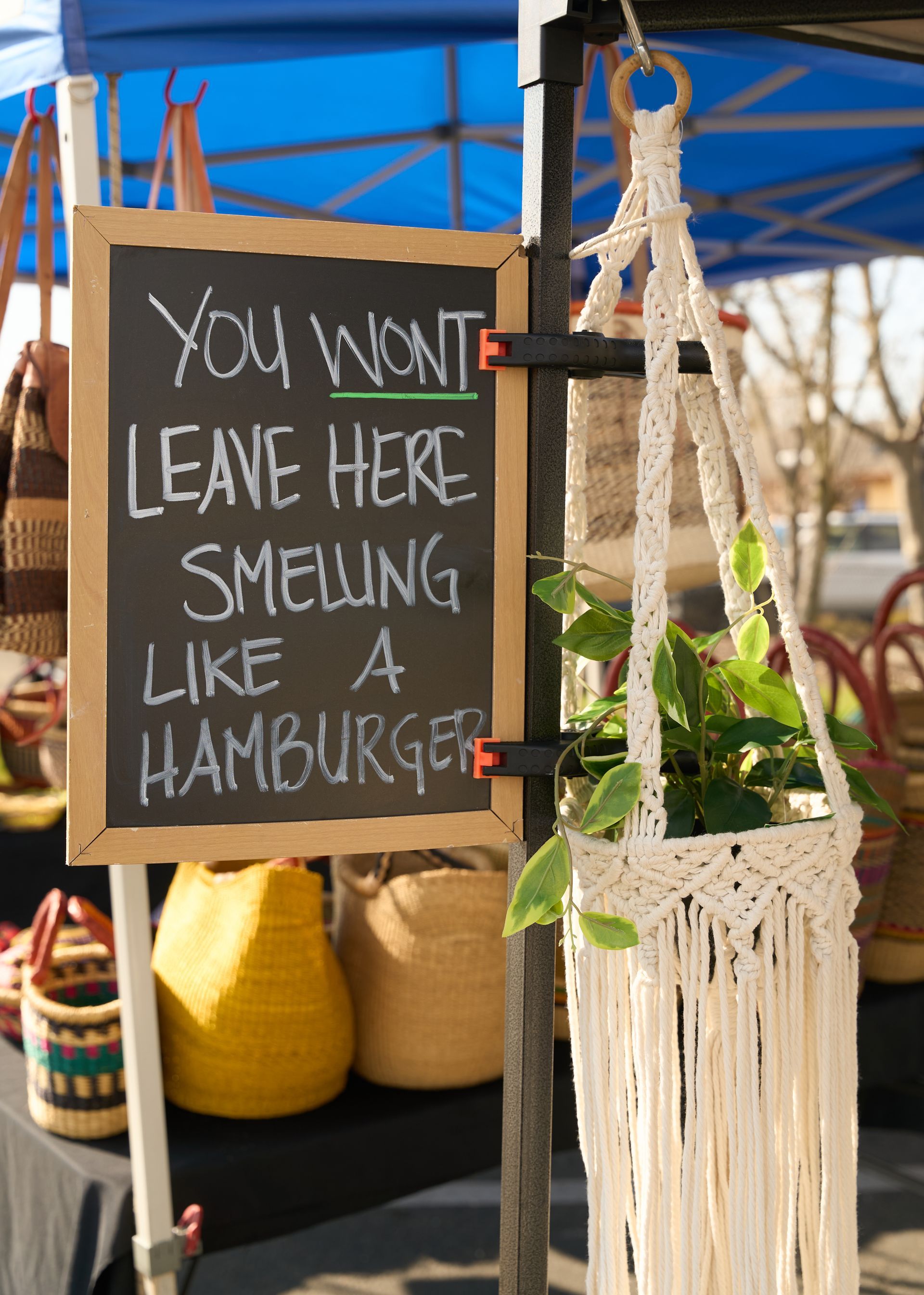 Handmade market sign reading 'You won't leave here smelling like a hamburger' displayed beside a hanging macramé plant hanger at the Loomis Community Farmers Market