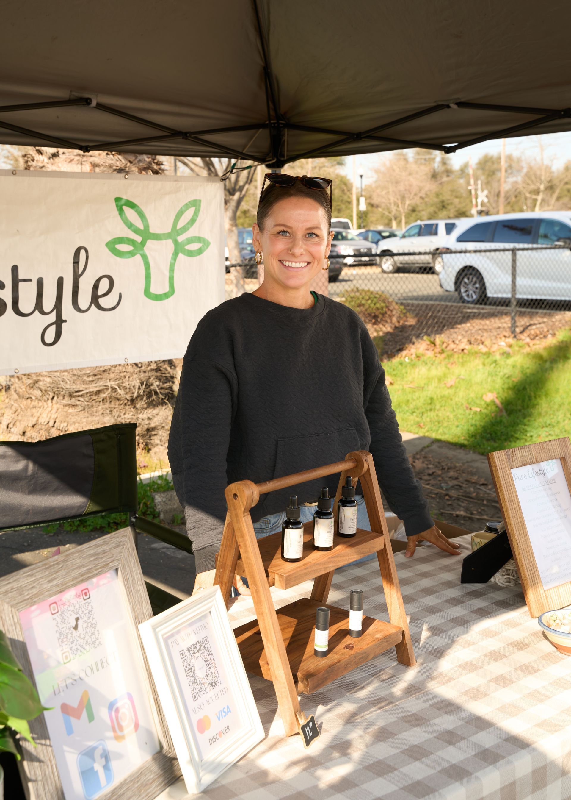 Smiling vendor standing behind a wooden display of artisan products at an outdoor canopy booth during the Loomis Community Farmers Market