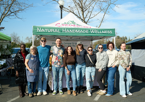 Group posing outdoors in front of a green Natural Handmade Wellness sign, with trees and blue sky behind them