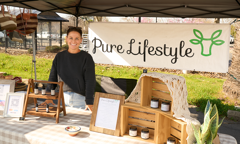 Vendor standing at an outdoor market stall under a canopy tent featuring a Pure Lifestyle banner and wooden display crates at the Loomis Community Farmers Market