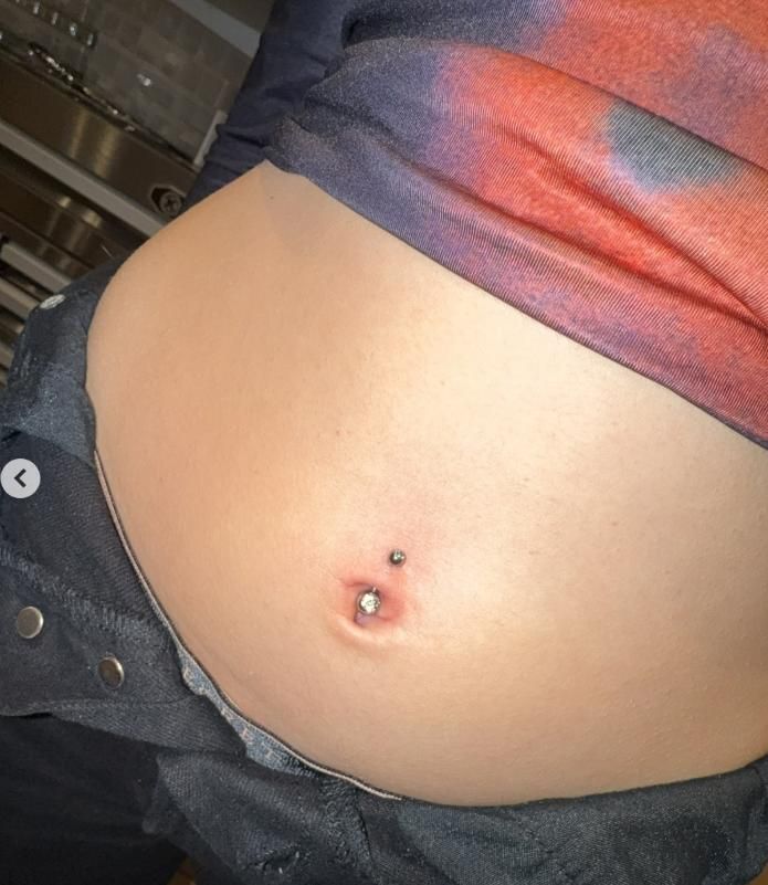 Person's abdomen with a navel piercing; wearing a tie-dye shirt and dark pants.