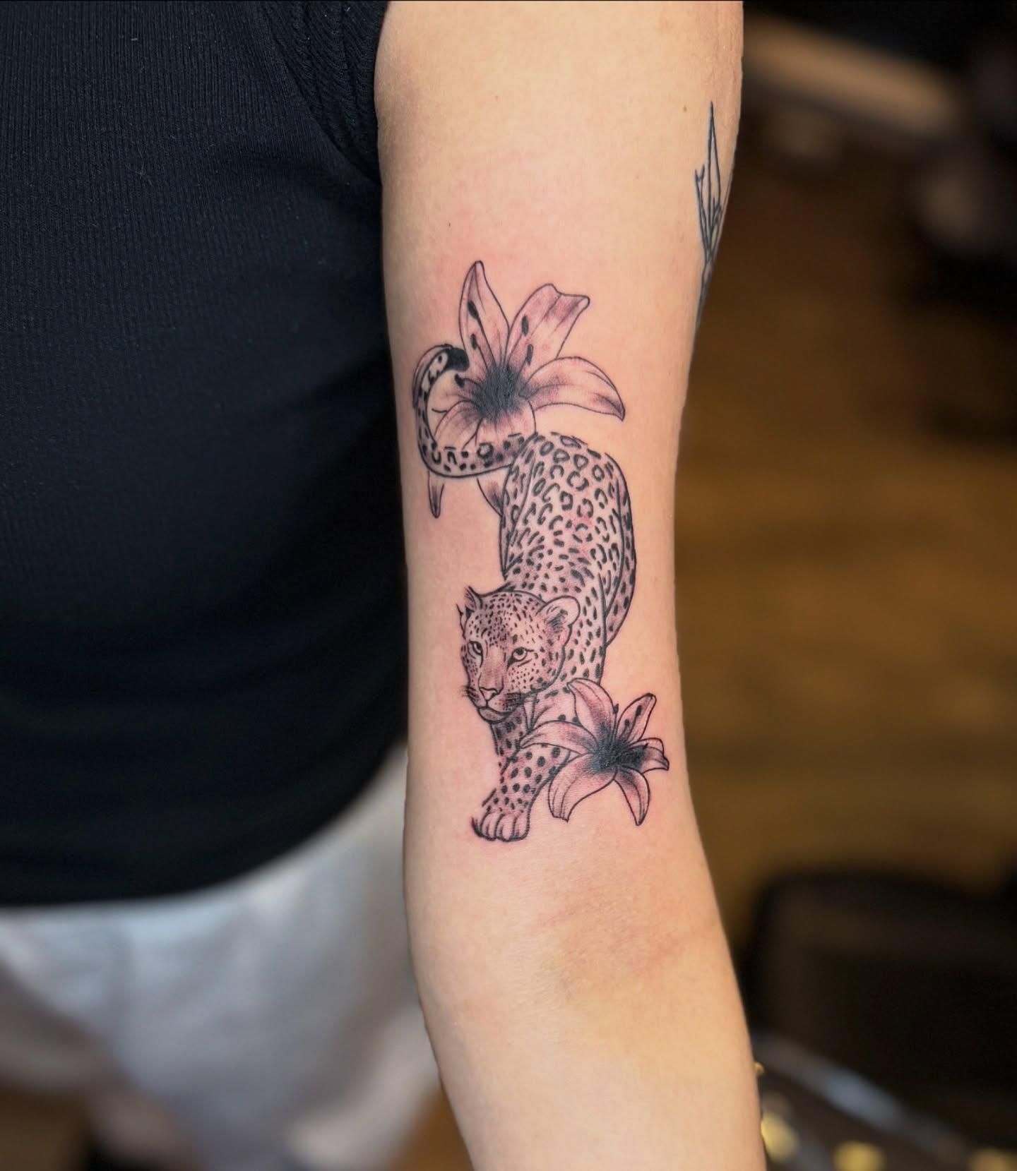 Tattoo of a leopard amidst lilies on an arm, black ink.