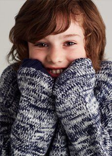 A young boy wearing a blue and white sweater is covering his face with his hands.