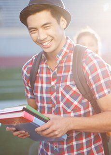 A young man in a plaid shirt is holding a stack of books and smiling.