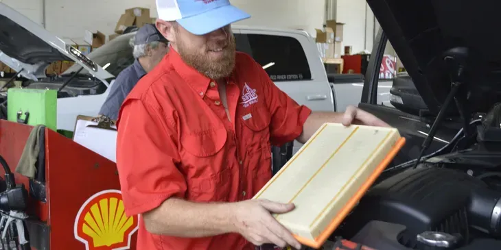 Mechanic replacing an air filter in a car engine bay. Man wearing red shirt, blue cap.