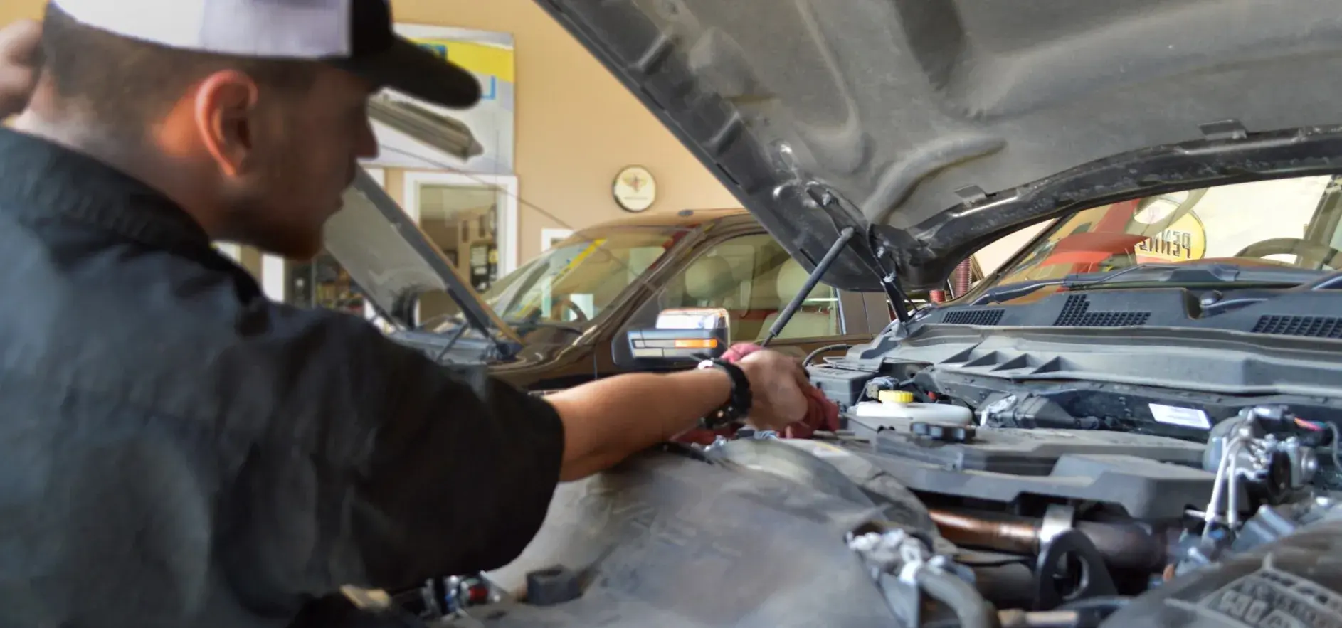 Mechanic works on a car engine in a garage. He wears a hat and is focused on his task.
