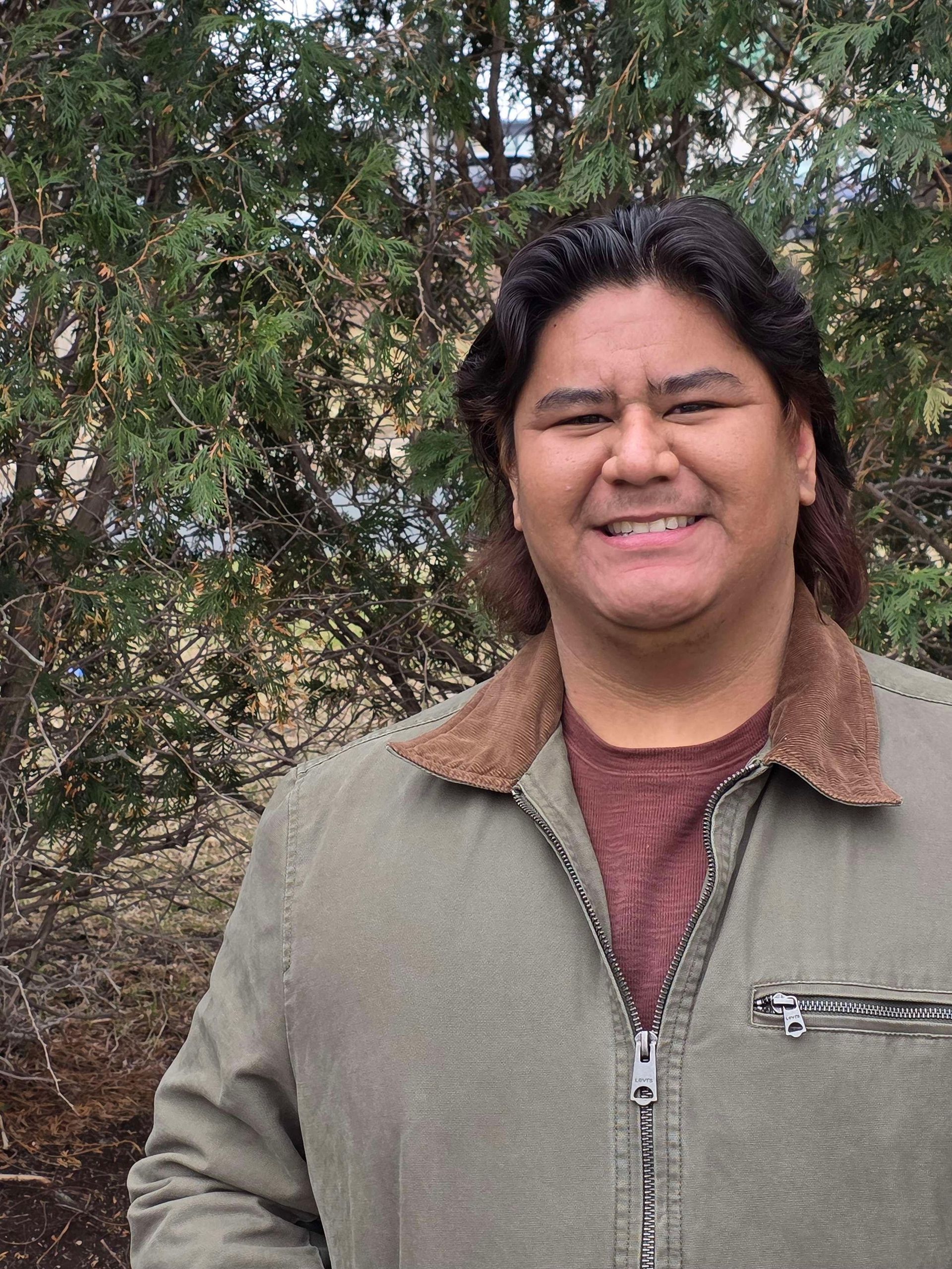 Man smiles outdoors, wearing a tan jacket with brown collar over a maroon shirt, against a backdrop of green foliage.